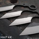 Four Bladetricks HAPAK karambit blades in progress on a steel bench, ground bevels bright against the dark forge finish, with the retention rings cut.