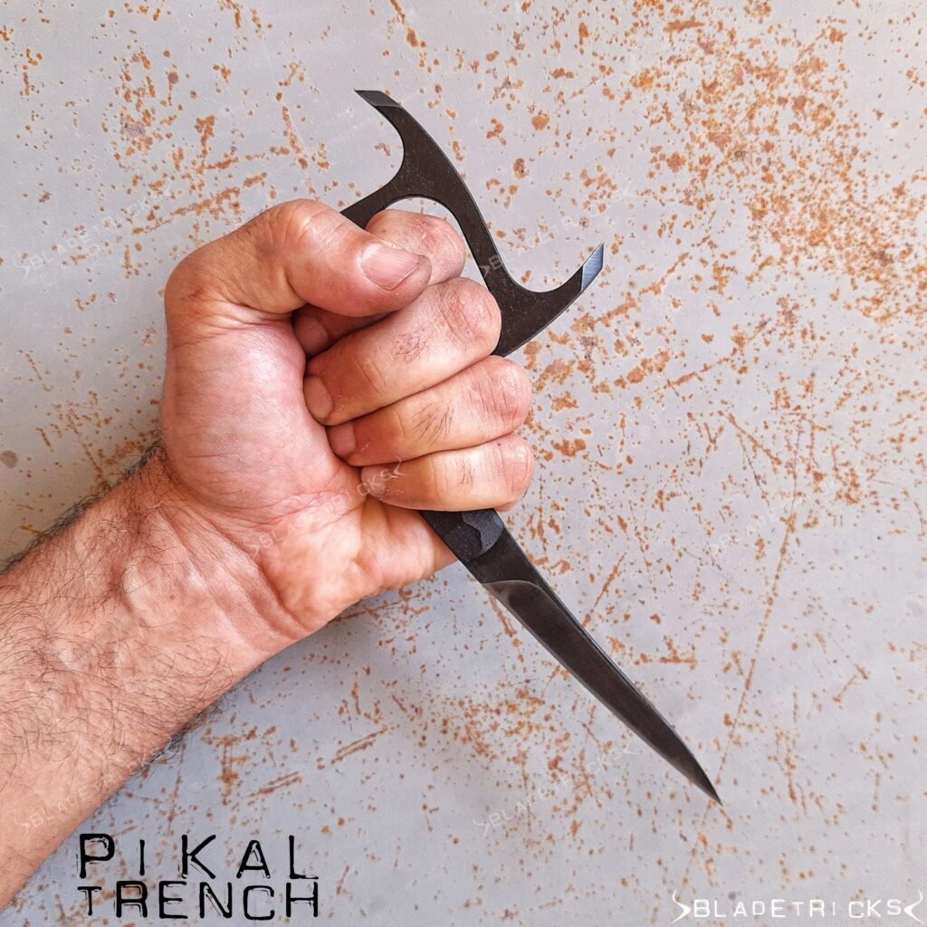 Bladetricks Pikal Trench held in a closed fist, the main blade running point-down below the hand and two horns projecting above it.