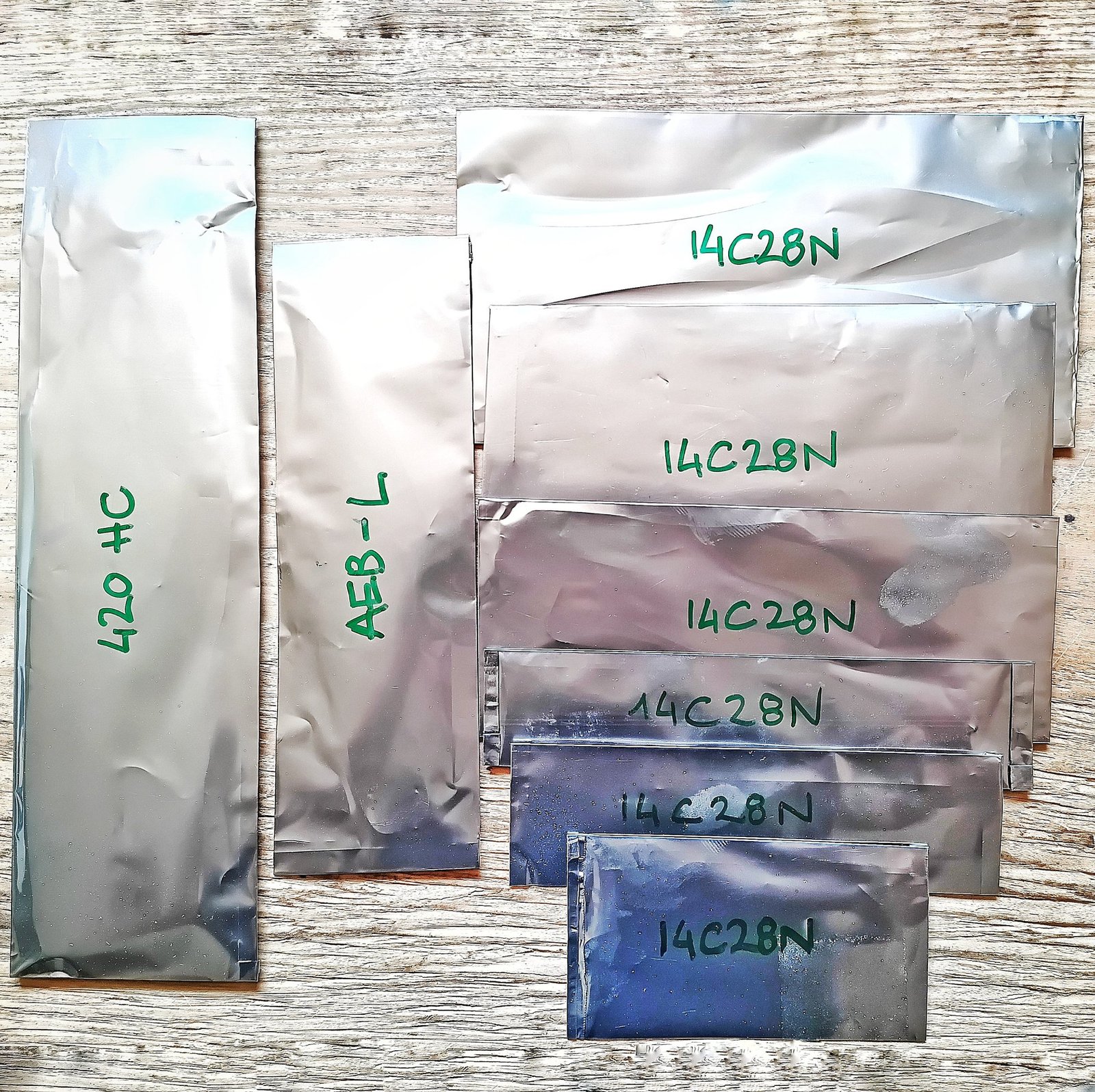 Stainless knife blades sealed in tool wrap foil, labeled 14C28N, AEB-L and 420HC, ready for the kiln